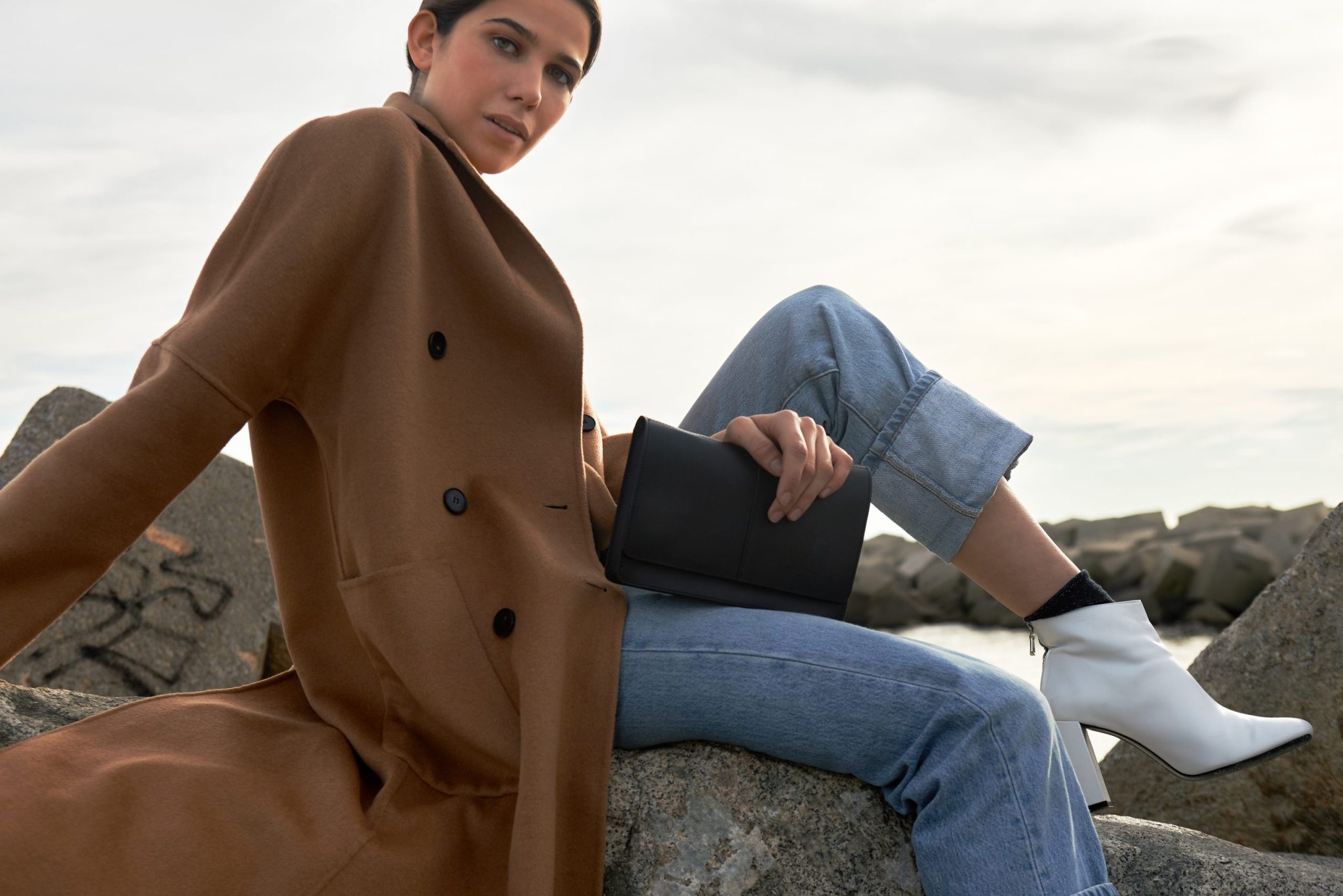 Boria&Coria: model in a brown coat with a black handbag sitting on rocks