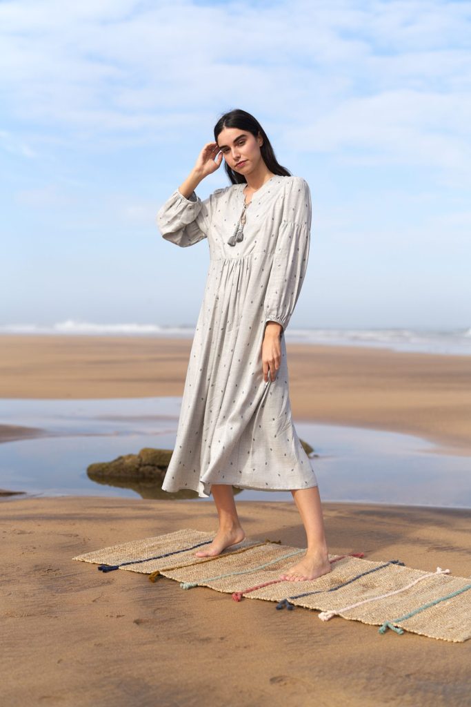 marwa: model in a polka-dot dress standing on a woven rug on the wet sand, with a tide pool behind