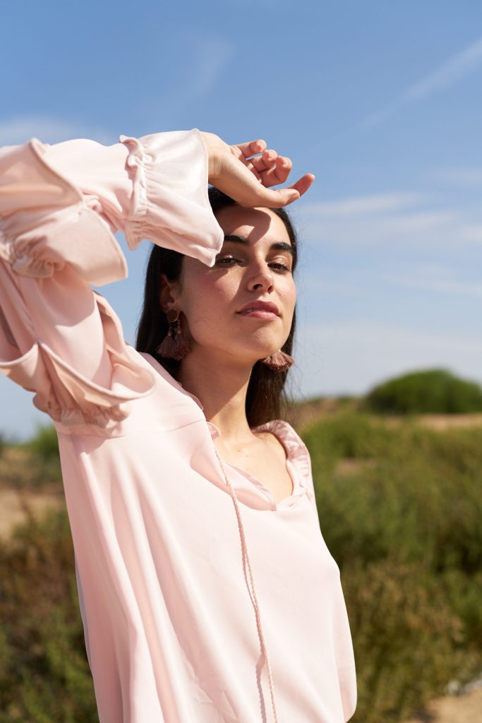 marwa: model in a pale pink blouse with her arm over her forehead in the sun, among shrubs