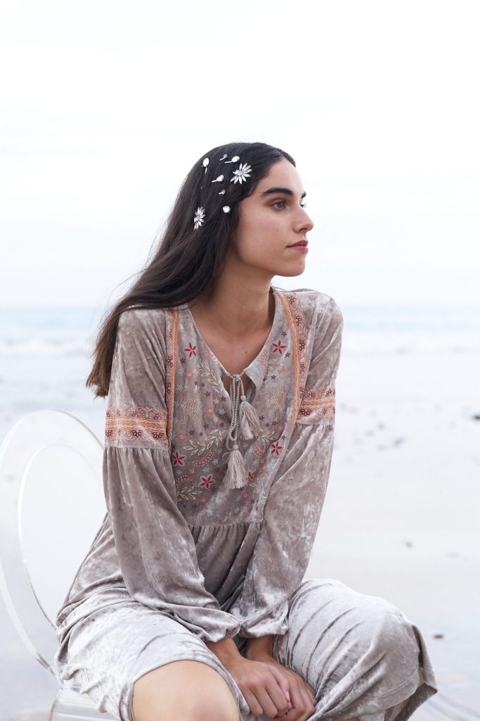 marwa: model in an embroidered velvet dress sitting in profile on a clear chair on the shore
