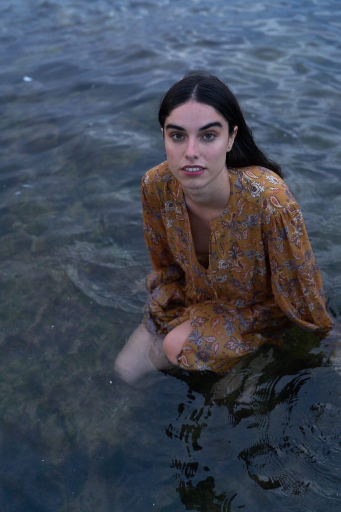 marwa: model crouching in the water in a mustard print dress, looking at the camera